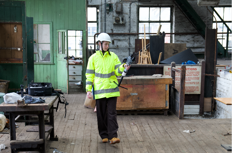 A surveyor wearing a white hard hat and high-visibility yellow jacket walks in an industrial building while holding a handheld SLAM laser scanner.