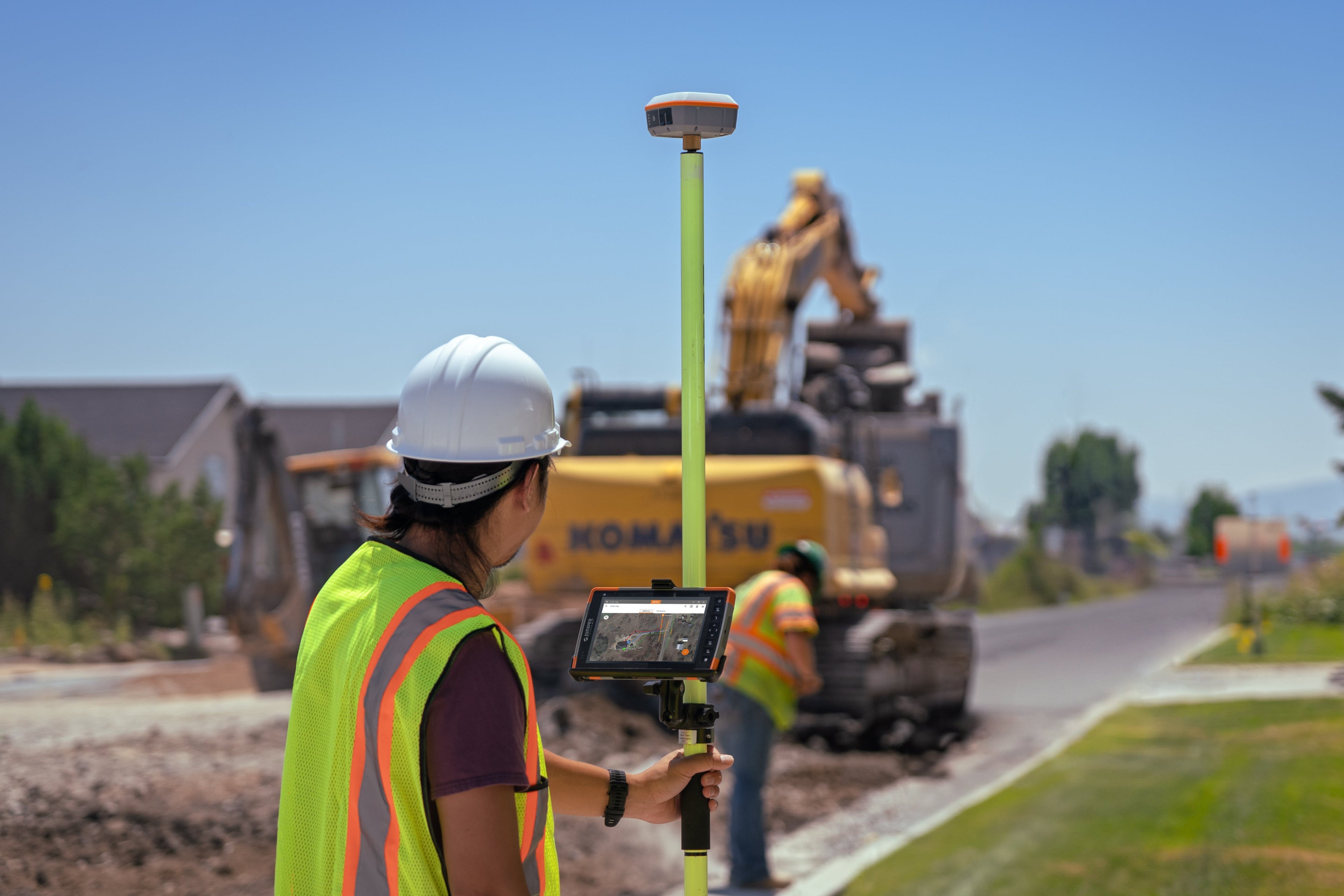 Person in safety gear using a Geode and Mesa Tablet on a construction site with equipment and trees in the background.